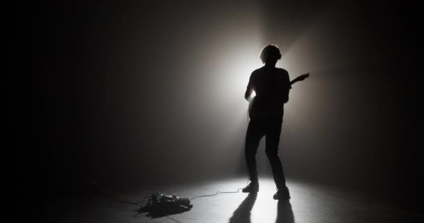 パフォーマンス中に輝く光と電気楽器を演奏するために暗闇に立っているエネルギッシュな男性ギタープレーヤーのリアルタイムフルボディ — ストック動画