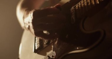 Yetişkin bir adam bej elektro gitarla müzik çalarken kafasını sallıyor ve loş ışıklandırmayla karanlık stüdyoda performans sergiliyor. 