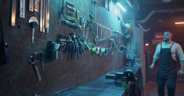ブルーネオンランプで照らされたガレージのツールウォールからレンチを取るエプロンのひげ付き男性労働者のショットでリアルタイムズーム — ストック動画