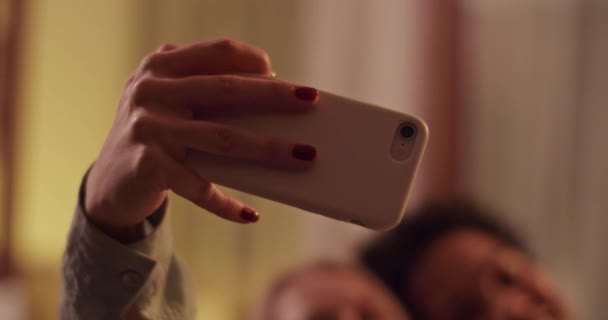 ハングアウト中にリビングルームでソーシャルネットワークのためにセルフィーを取っている間 スマートフォンを使用して若い多民族幸せな女性のラックフォーカスハンドヘルドショット — ストック動画