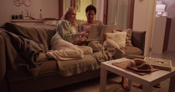 夜の快適なソファーで休憩し 週末に一緒に写真アルバムを見ている若い多人種間のガールフレンドのリアルタイムハンドヘルドショット — ストック動画