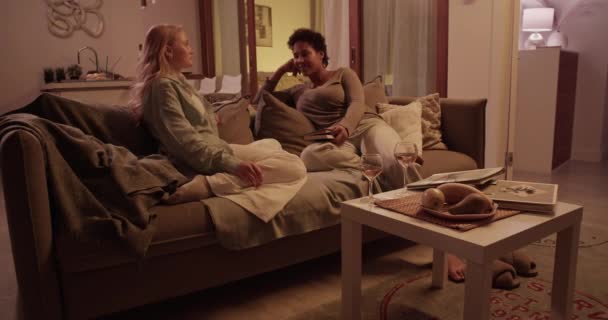 若い幸せなアフリカ系アメリカ人女性のリアルタイムフルボディは コーヒーテーブルからワイングラスを取っているガールフレンドとチャットしながら手を傾ける本とソファに座っています — ストック動画