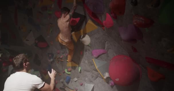 ジムでのボルダリングトレーニング中に壁に登るシャツレスの男性アスリートを導くトレーラーのハンドヘルドショット — ストック動画