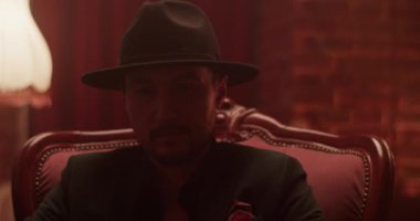 Gerçek zamanlı olarak el kamerasında, parlak lamba ve tuğla duvara karşı karanlık bir odada minderli koltukta otururken kendine güvenen sakallı Meksikalı bir erkek el feneriyle yukarı bakıyor.