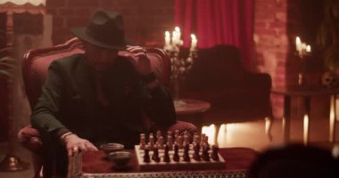 Takım elbiseli ve şapkalı kendine güvenen yetişkin bir adam evdeki karanlık odada satranç oynuyor.