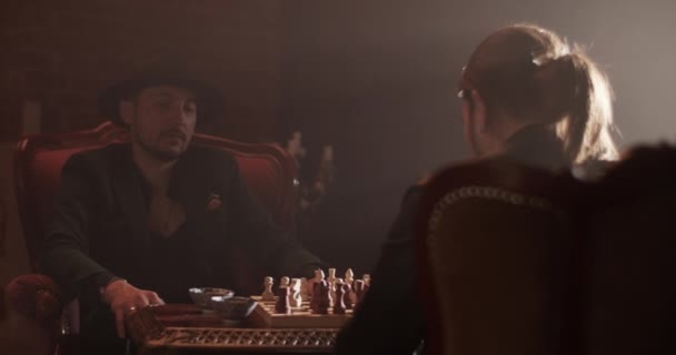 自宅のダークルームで匿名の友人とチェスボードゲームをするのに帽子とスーツの自信を持った大人の男 — ストック動画