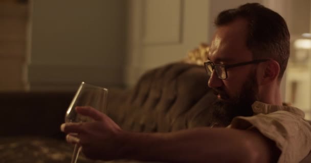 ヴィンテージハウスの快適なソファーでリラックスしながらガラスからワインを飲む眼鏡の大人のひげ付き男のハンドヘルドショット — ストック動画
