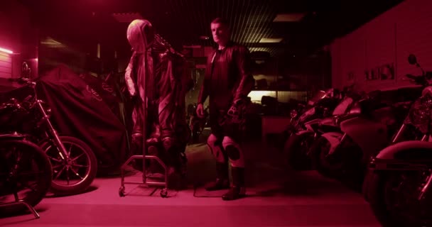 赤いネオン ライトを輝かせる暗いガレージでレースのために準備している間特別な衣装を身に着けている専門の男性のオートバイのリアルタイムハンドヘルドショット — ストック動画