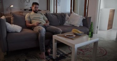 Yalnız sakallı yetişkin bir adam evdeki oturma odasında kanepede uzanırken uzaktan kumandayla televizyon kanallarını değiştiriyor.