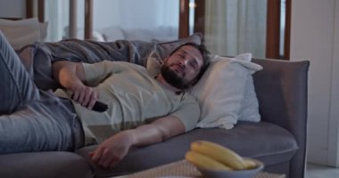 Gerçek zamanlı sakallı, tembel, yetişkin bir erkek uzaktan kumandayla kanepede uzanıyor ve bulanık camlı pencerelerle odada zaman geçiriyor.