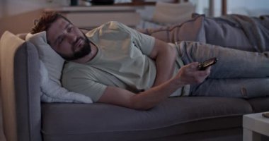 Evde oturma odasındaki kanepede uzanırken uzaktan kumandayla sık sık televizyon kanallarını değiştiren gündelik giysiler içinde yalnız sakallı yetişkin bir erkek.