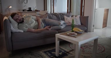 Depresyondaki sakallı yetişkin adam evdeki oturma odasında kanepede uzanırken uzaktan kumandayla televizyon kanallarını değiştiriyor.