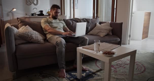 疲れた大人の男性は ノートパソコンを閉じるソファの上に座って 自宅のリビングルームにテーブルの上に置くカジュアルな服で — ストック動画
