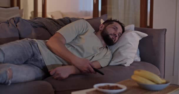 カジュアルな服の孤独な成人男性は リモートコントロールを使用して頻繁にテレビチャンネルを変更し 自宅のリビングルームのソファーに横たわる — ストック動画