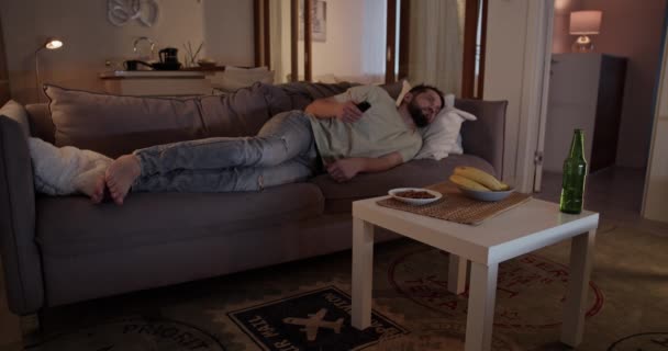 穿着休闲装的懒汉在家里客厅的沙发上躺着用遥控器换电视频道 — 图库视频影像
