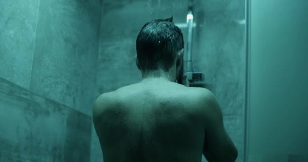 シャワーの下に立っている間 そして光の下の浴室で顔に手で水を拭いている間 匿名のシャツレスの落ち込んだ男性のリアルタイムのバックビュー — ストック動画