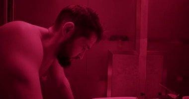 Banyoda aynaya bakan sakallı, depresif, üstsüz, yetişkin bir adamın sağa dön. Kırmızı ışıkla aydınlatılmış.