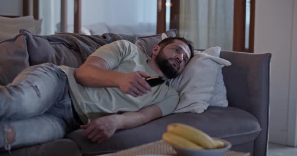 穿着休闲装的孤独成年男子在家客厅里躺在沙发上 经常用遥控装置更换电视频道 — 图库视频影像