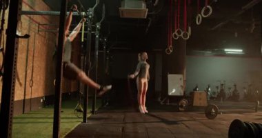 Barda barfiks çeken kaslı bir adamın gerçek zamanlı yan görüntüsü ve spor salonunun halı zemininde kadın halı atlama halatı ve ekipmanlarıyla antrenman yapması.
