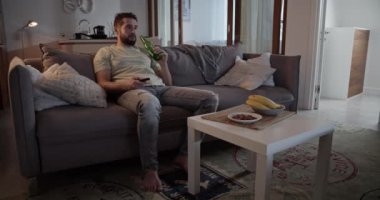 Depresyondaki sakallı yetişkin adam elinde uzaktan kumandayla evde oturma odasındaki kanepede oturup bira içerken televizyon izliyor.