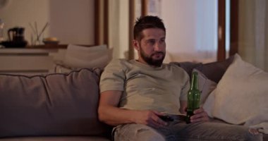 Gerçek zamanlı mutsuz yetişkin sakallı erkek uzaktan kumandayla kanepeye uzanıp evde televizyon izlerken biradan bira içerken başka tarafa bakıyor.