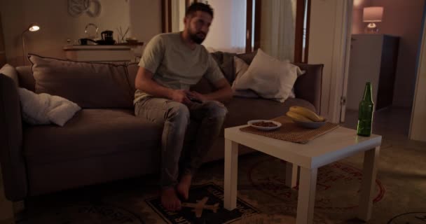 自宅のリビングルームでソファーに横たわっている間 リモートコントロールを使用してテレビチャンネルを変更するカジュアルな服の孤独な大人の男 — ストック動画