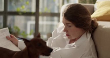 Gerçek zamanlı yetişkin bir kadın, elinde dizüstü bilgisayarla yere yatıp çalışırken belgeye bakıyor ve evde öpüşürken kalemle köpek burnunu okşuyor.