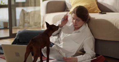 Gerçek zamanlı yetişkin bir kadın, elinde dizüstü bilgisayarla yere yatıp çalışırken belgeye bakıyor ve evde öpüşürken kalemle köpek burnunu okşuyor.