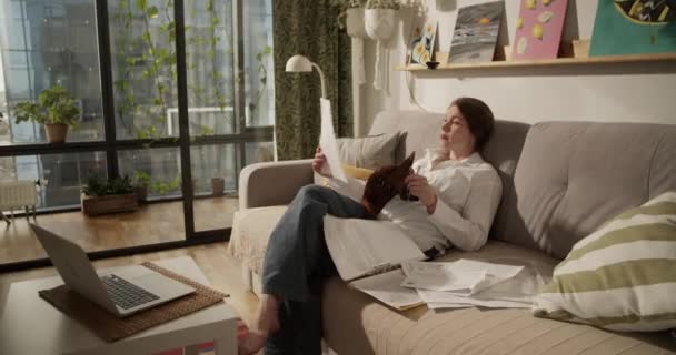 実時間のハンドヘルドショット 裸足の大人の女性読書文書ファイルの側面のビューは 家からの仕事中に犬とラップトップとソファーに横たわって座っている間 — ストック動画