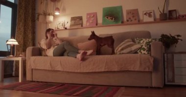 Çıplak ayaklı yetişkin bir kadının gerçek zamanlı yan görüntüsü kanepeye uzanmış cilt bakım maskesi takıyor ve akşam vakti oturma odasında köpekleri okşuyor.