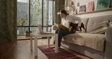 Gerçek zamanlı olarak çıplak ayaklı yetişkin bir kadının laptopta çalışırken gözlerini kaçırması belgelere atıfta bulunması ve kanepede evcil köpekle oynaması. 