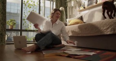 Gerçek zamanlı gülümseyen, çıplak ayaklı yetişkin bir kadın, evde çalışırken dizüstü bilgisayarla yerde oturup belgeyi kontrol ederken köpekten kalem topluyor.