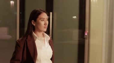 Gece aydınlık ışıklarda modern binanın cam duvarları boyunca yürürken şık takım elbiseli genç Asyalı kadın modelin gerçek zamanlı fotoğrafını izliyorum.