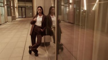 Modern takım elbiseli, havalı, genç Asyalı kadın modelin omuz omuza, elleri belde ve cebinde cam duvara yaslanırken kameraya bakışını yakınlaştır.