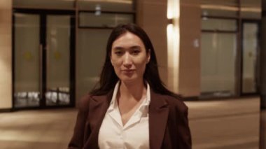 Gerçek zamanlı olarak gülümseyen Asyalı, ceketli, cam duvarların yanındaki ışıklarla aydınlatılmış şehrin avlusunda yürürken kameraya bakan genç kadın manken portresinin izini sürüyorum.