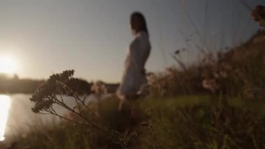Güneşli yaz gününde bulutsuz gökyüzünün altında sakin göl kenarında, çimenli bir arazide duran modaya uygun beyaz elbiseli genç bir kadının yan bakışını çek.