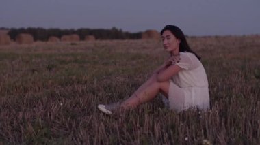 Gün batımında kırsalda oturan beyaz elbiseli mutlu Asyalı kadın modelin el kamerasıyla tüm vücudu görüntülendi.