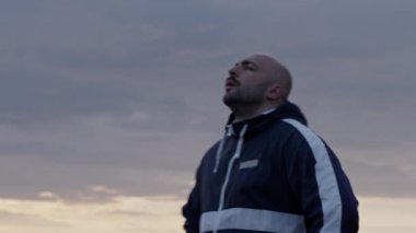 Kapüşonlu ceketli yetişkin erkek sporcunun gün batımında bulutlu gökyüzü altında yoğun idmandan sonra derin derin nefes alışları.