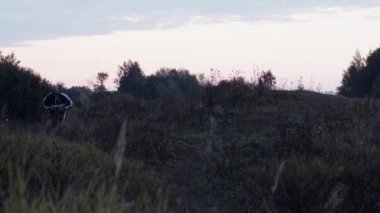 Gün batımında bulutlu gökyüzüne karşı yeşil bitkilerle tepelik arazide koşarken el kamerasıyla gerçek zamanlı yetişkin erkek sporcu bakışları.