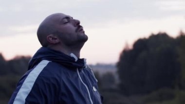 Gerçek zamanlı fotoğraf, yorgun yetişkin erkek koşucu gün batımında nefes alırken yeşil ağaçlarla bulanık doğaya karşı uzaklara bakıyor.