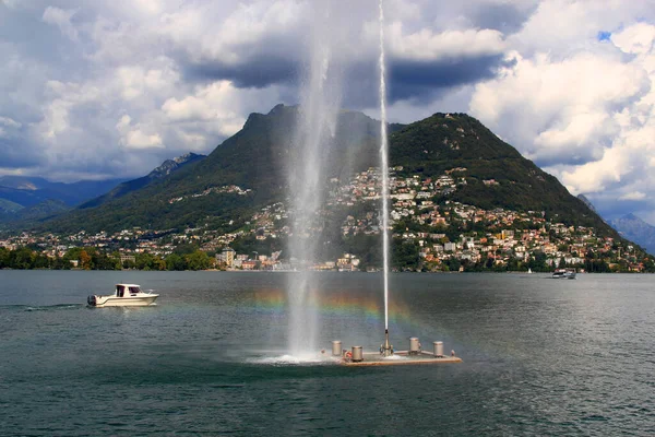 Landscape with a view of the mountains, a boat on Lake Lugano, with a fountain and a rainbow in the foreground against a stormy sky, in the city of Lugano in southern Switzerland