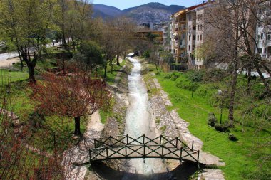 A view of a park area with a river and an iron bridge across it, and the yellow Setbasi Koprusu bridge and a mountain in the background in the city of Bursa, Turkiye