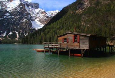 Lago di Braies Gölü 'nün zümrüt düz yüzeyi, önünde bir kulübe ve arkasında kar örtülü dağlar, Dolomitler, İtalya' da bulutlu mavi bir gökyüzüne karşı.