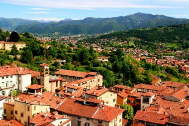 Bergamo şehrinin (İtalya) bir kısmının manzarası olan şehir, arka planında bulutlu mavi bir gökyüzüne karşı yeşil dağlar yer alıyor.