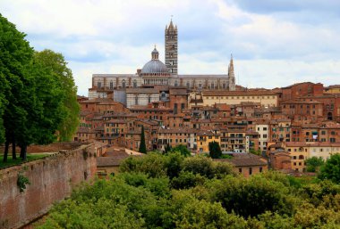 Siena şehrinin tarihi kısmının panoramik manzarası Duomo di Siena ve ön planda yeşil ağaçlar ve çalılar İtalya 'nın Toskana bölgesinde bulutlu mavi bir gökyüzüne karşı