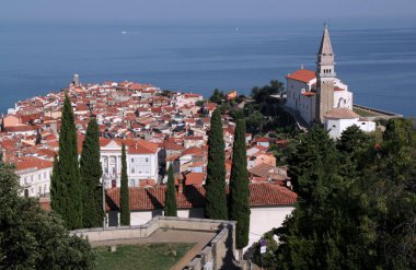 Arkasında tarihi şehir merkezi, kilise, deniz feneri, selvi ağaçları ve Adriyatik Denizi bulunan Piran (Slovenya) şehri manzarası