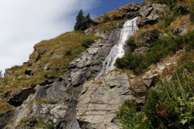 Romanya 'daki Transfagarasan dağ yolunda bulutlu mavi gökyüzüne karşı yüksek bir uçurumdan düşen şelale manzaralı manzara fotoğrafı