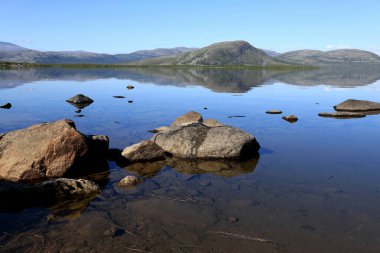 Tornetrask Gölü 'nün düz ayna yüzeyine bakan manzara fotoğrafı ön planda büyük taşlar ve arka planda Abisko yakınlarında bir dağ sırası, kuzey İsveç