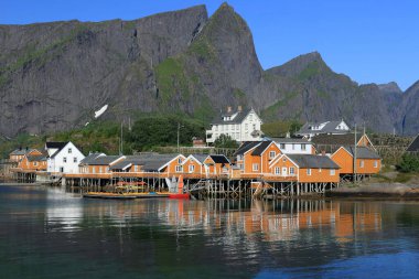 Norveç 'in Lofoten Adaları' ndaki Sakrisoya Adası 'ndaki suya yansıyan dağların ve sarı rorbu evlerinin manzaralı fotoğrafı.
