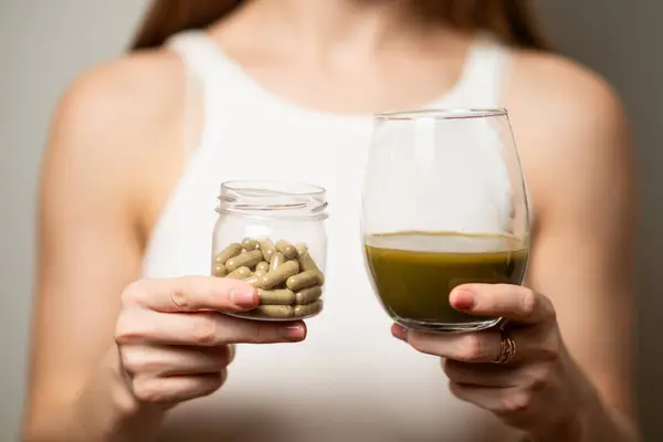 Kratom tüketim konsepti: dişi, bir bardak yeşil sıvı içmeye hazır, sıcak suda eritilmiş kratom tozu, bir bardak çay ve kavanozda bir avuç hap, yakın çekim fotoğrafı, seçici odaklanma.